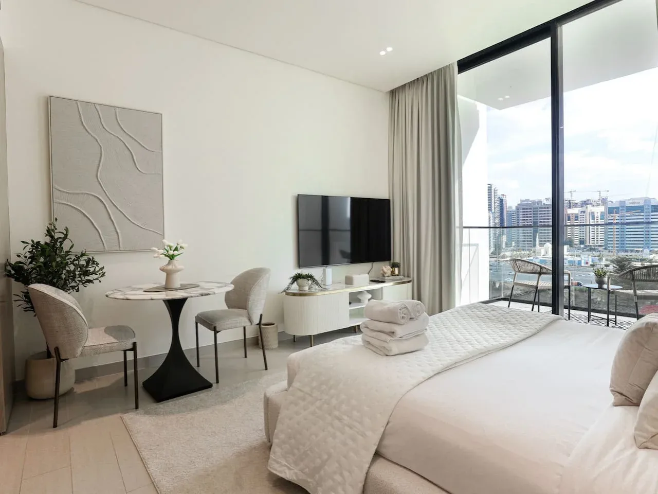 A bright bedroom with a neatly made bed, seating area, and large windows overlooking the city.