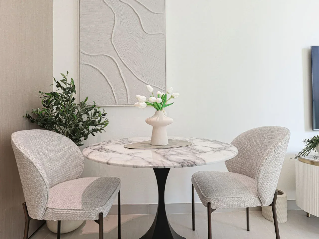 A small dining area has a round table, two upholstered chairs, and a vase of flowers.