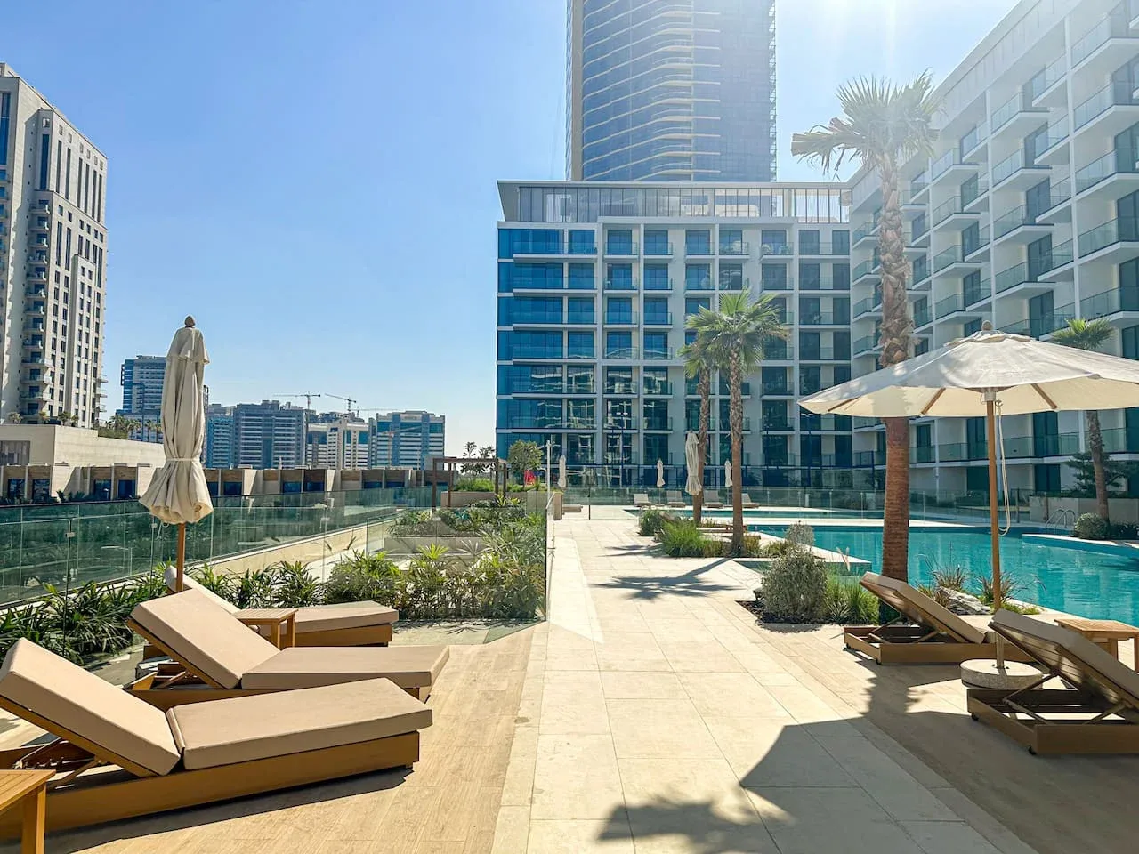A sunny outdoor pool area with lounge seating, umbrellas, and modern high-rise buildings.