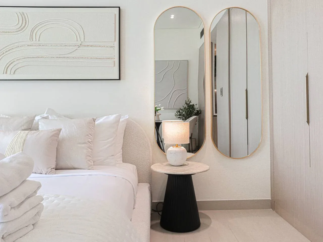 A bright bedroom with a neatly made bed and two arched mirrors.