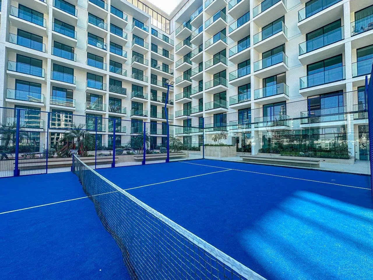A blue tennis court with a net sits in the courtyard of a modern building.