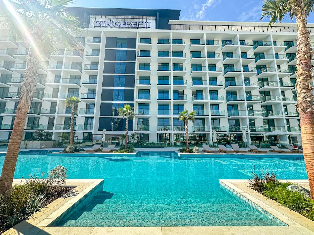 A hotel building overlooks a turquoise swimming pool lined with palm trees.