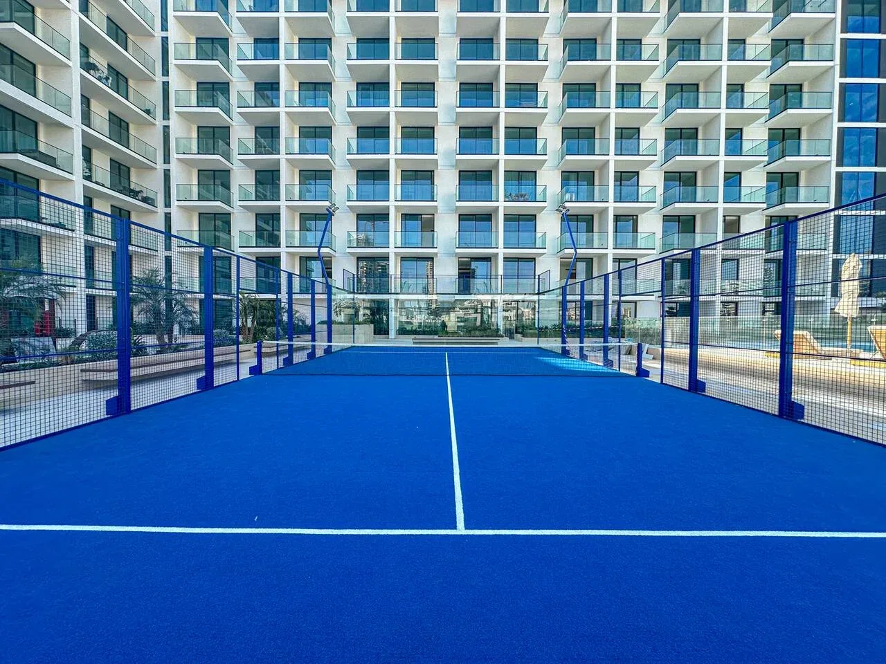 A blue padel court sits in front of a modern building with balconies.