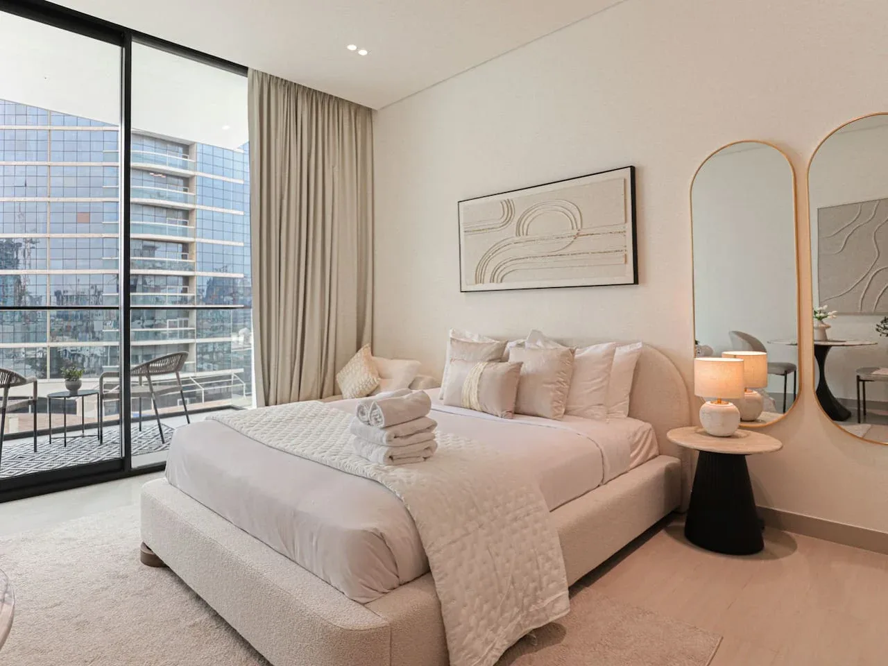 A bright bedroom with a neatly made bed beside floor-to-ceiling windows.