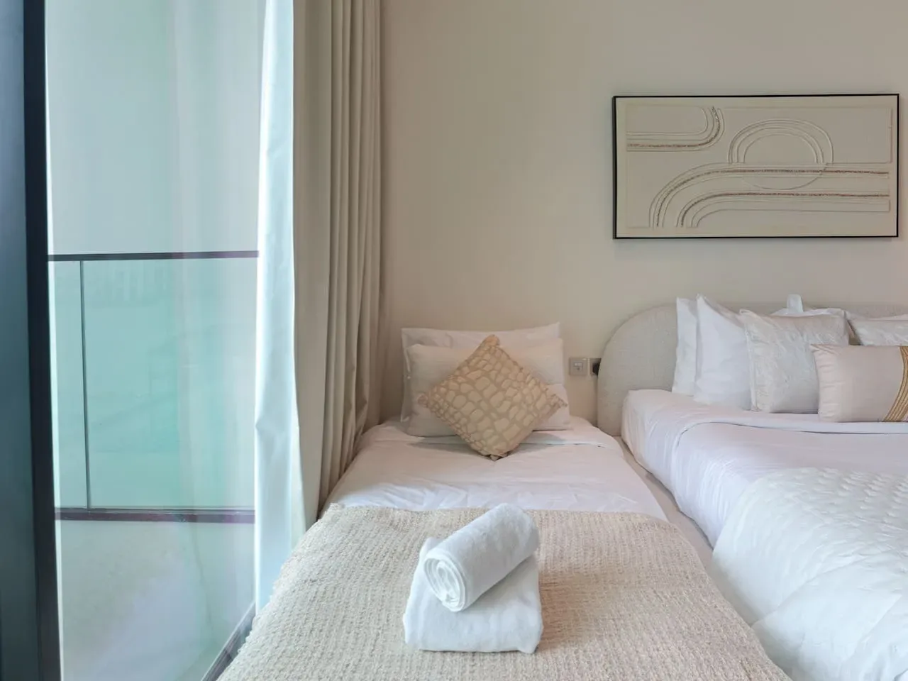 A bright bedroom with two beds, pale bedding, and a framed wall print.