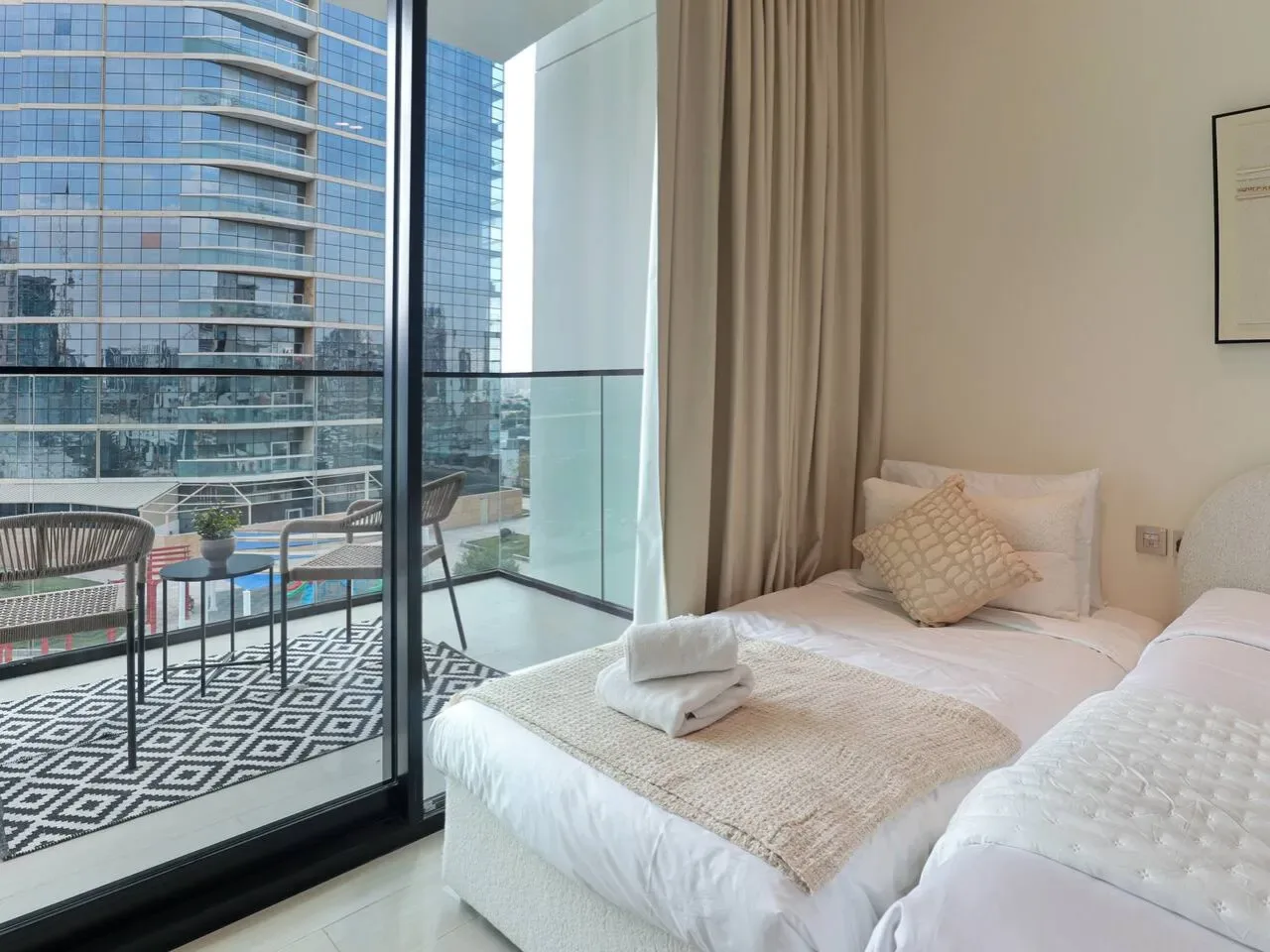 A bright bedroom with a bed beside floor-to-ceiling windows overlooking nearby buildings.