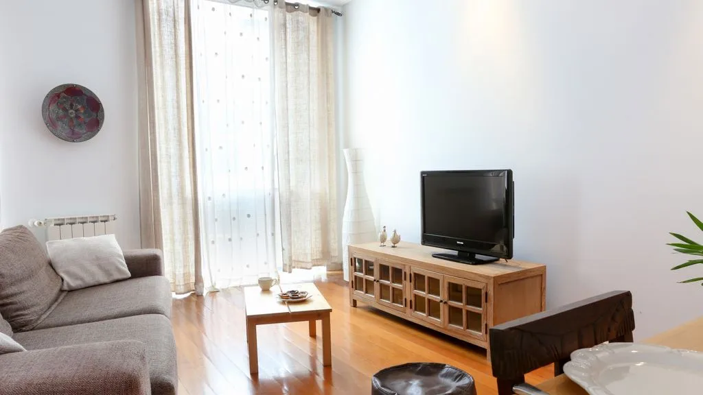 Una sala luminosa con asientos, un televisor y ventanas altas con cortinas.