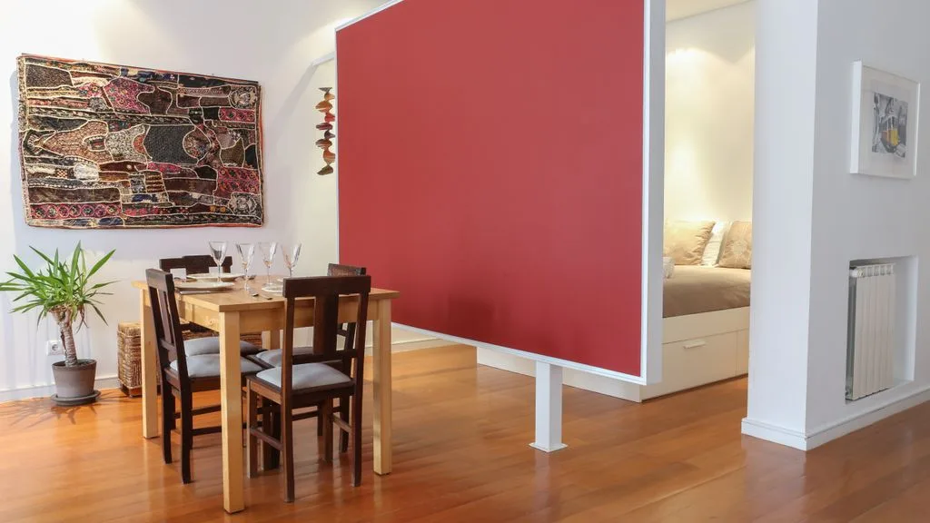 Comedor con asientos de madera junto a un separador rojo y un dormitorio contiguo.