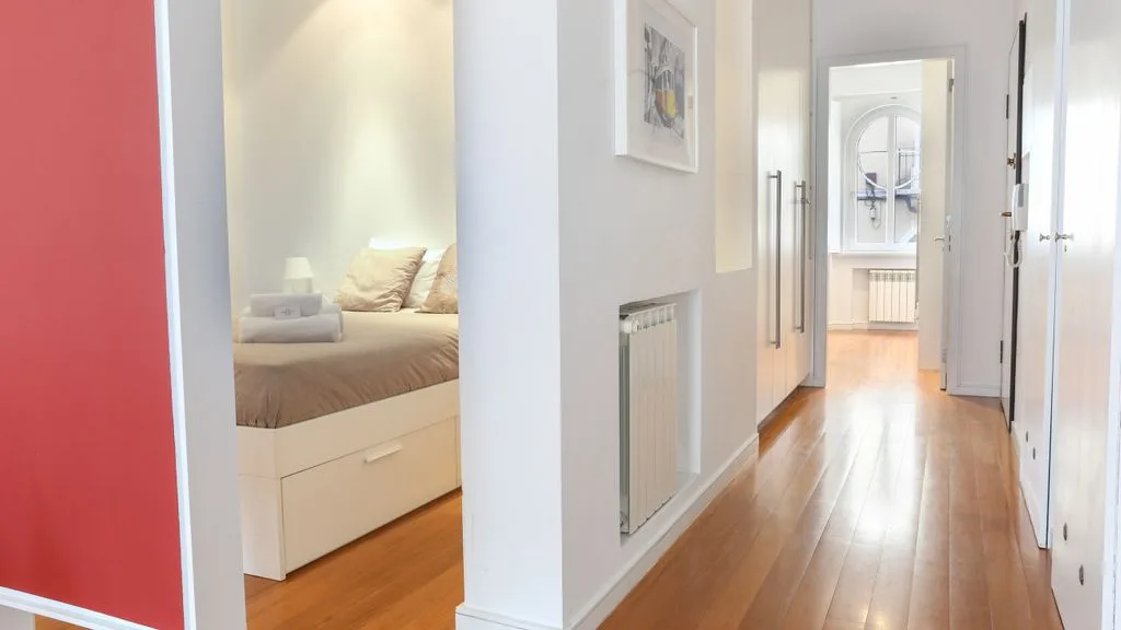 Un pasillo luminoso con suelo de madera conduce hacia un dormitorio y una ventana.