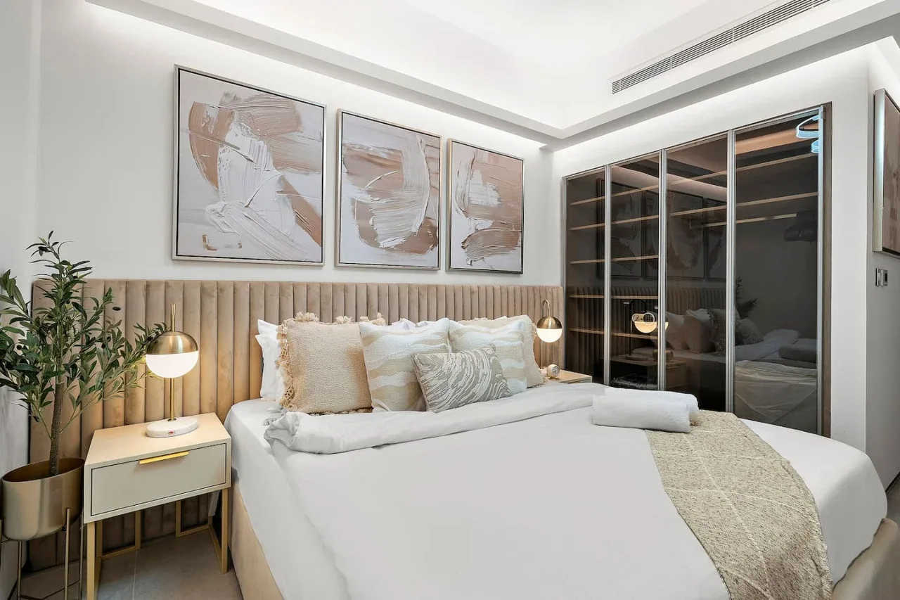Un dormitorio luminoso con una cama tendida y cuadros sobre el cabecero.