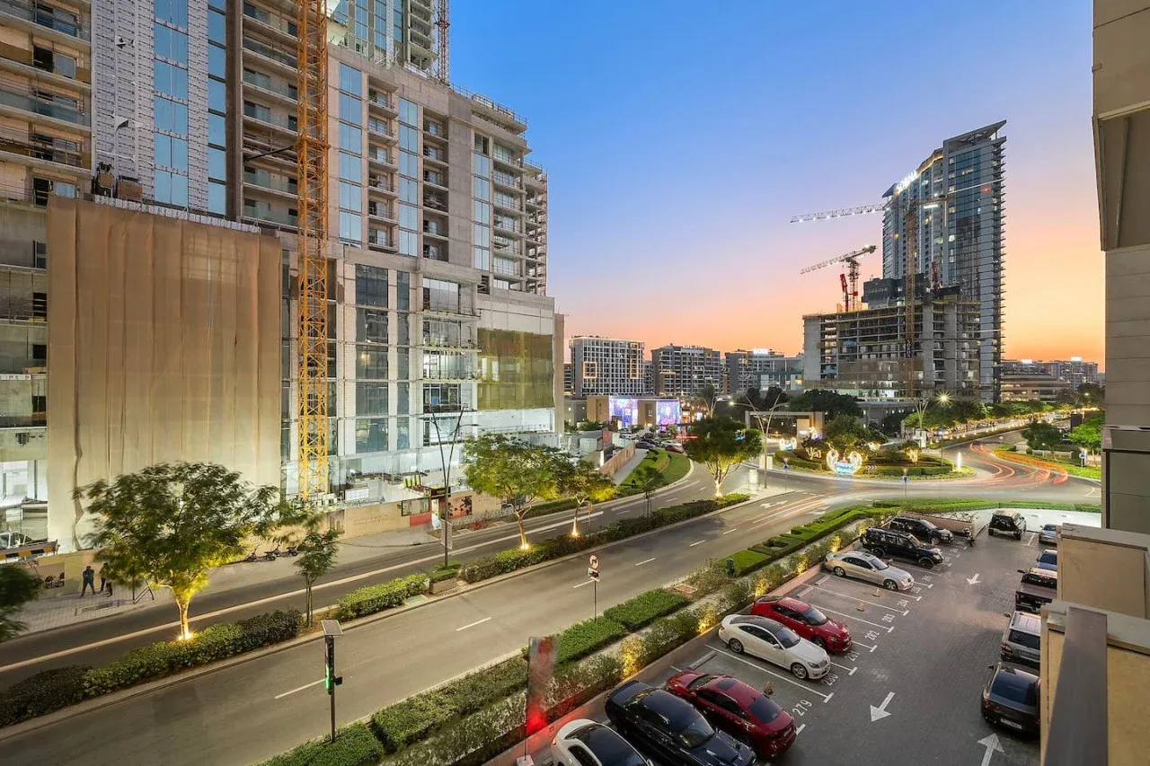 Una calle urbana y una zona de estacionamiento están rodeadas de edificios altos al atardecer.