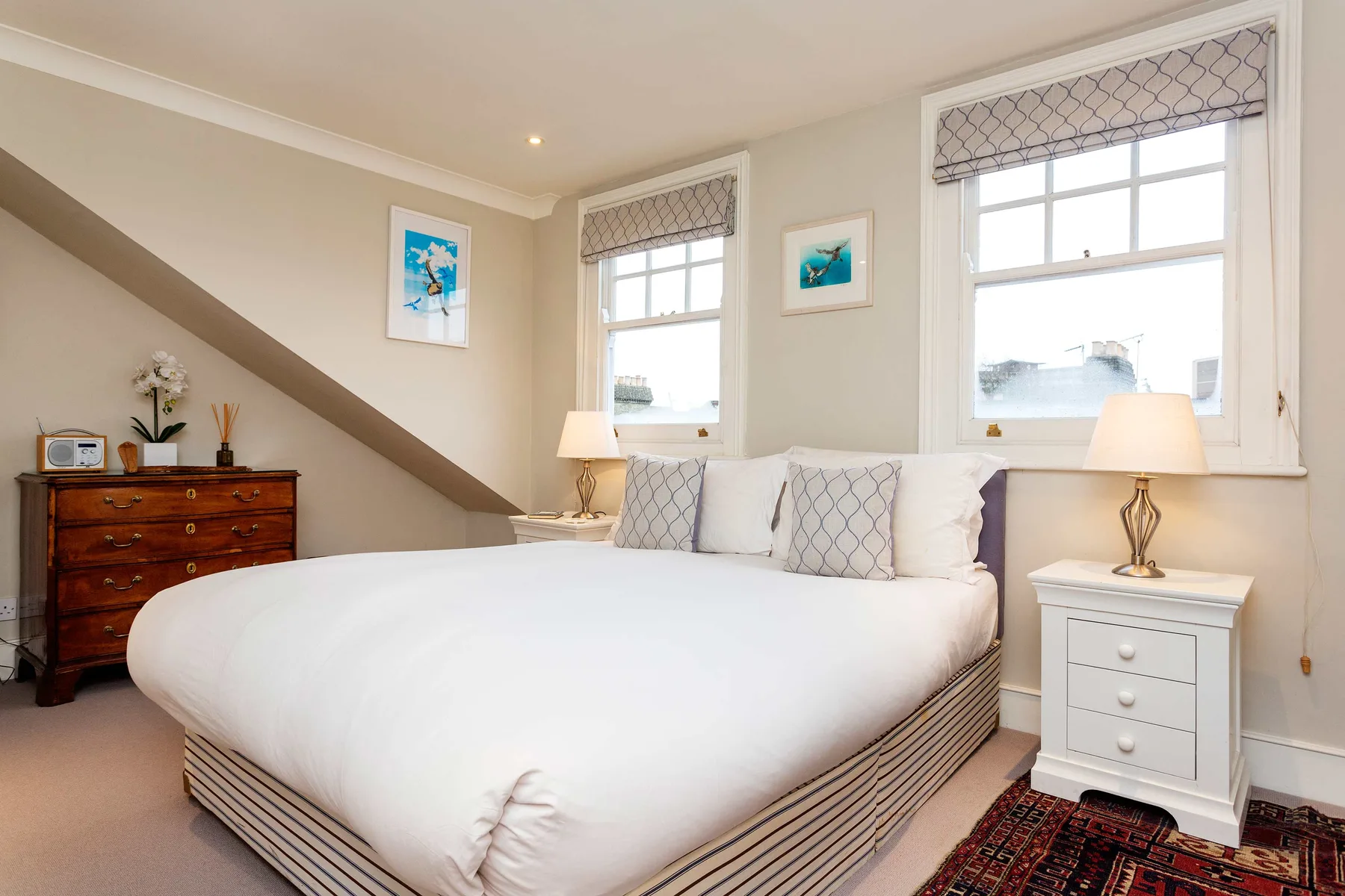 A bright bedroom with a large bed, bedside table, and two windows.