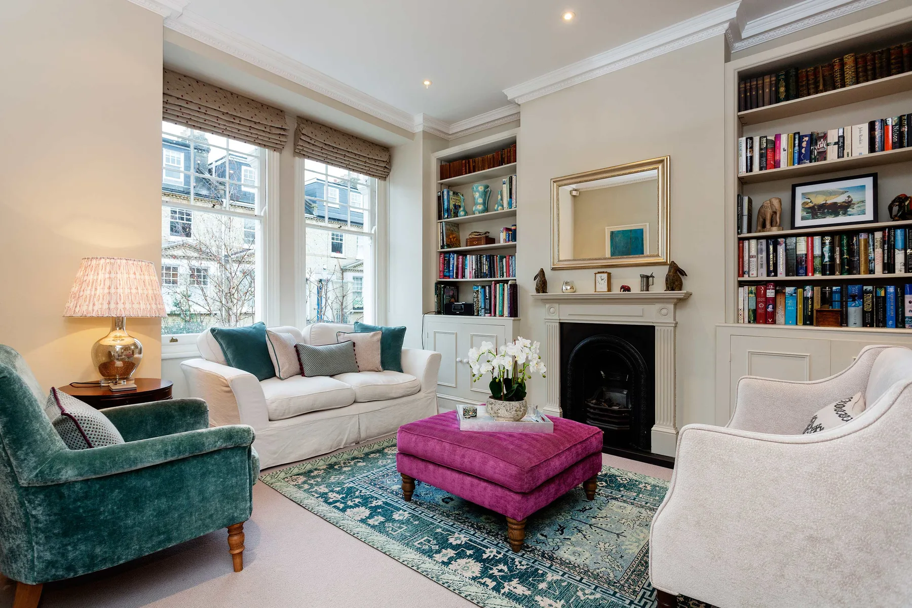 A bright living room with seating around a fireplace and a colorful ottoman.