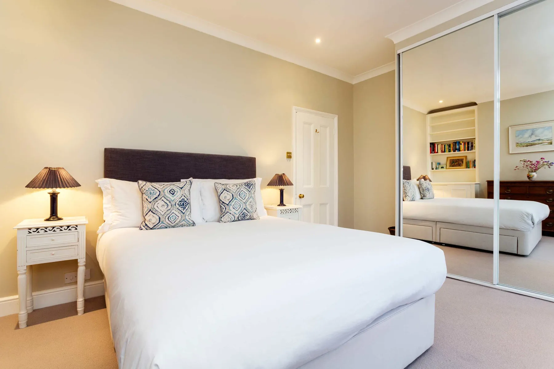 A bedroom with a neatly made bed, bedside tables, and mirrored wardrobe doors.