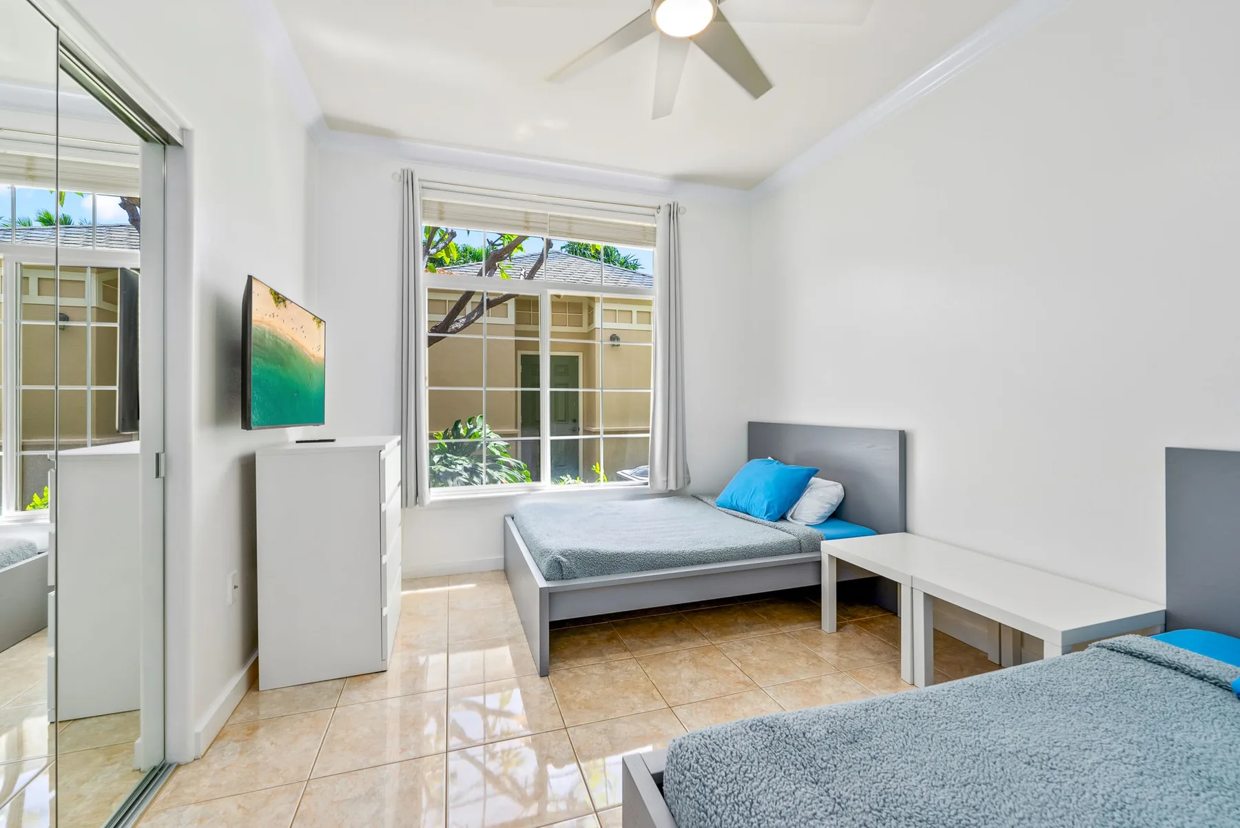 Un dormitorio luminoso con dos camas, un ventilador de techo y ventanas grandes.