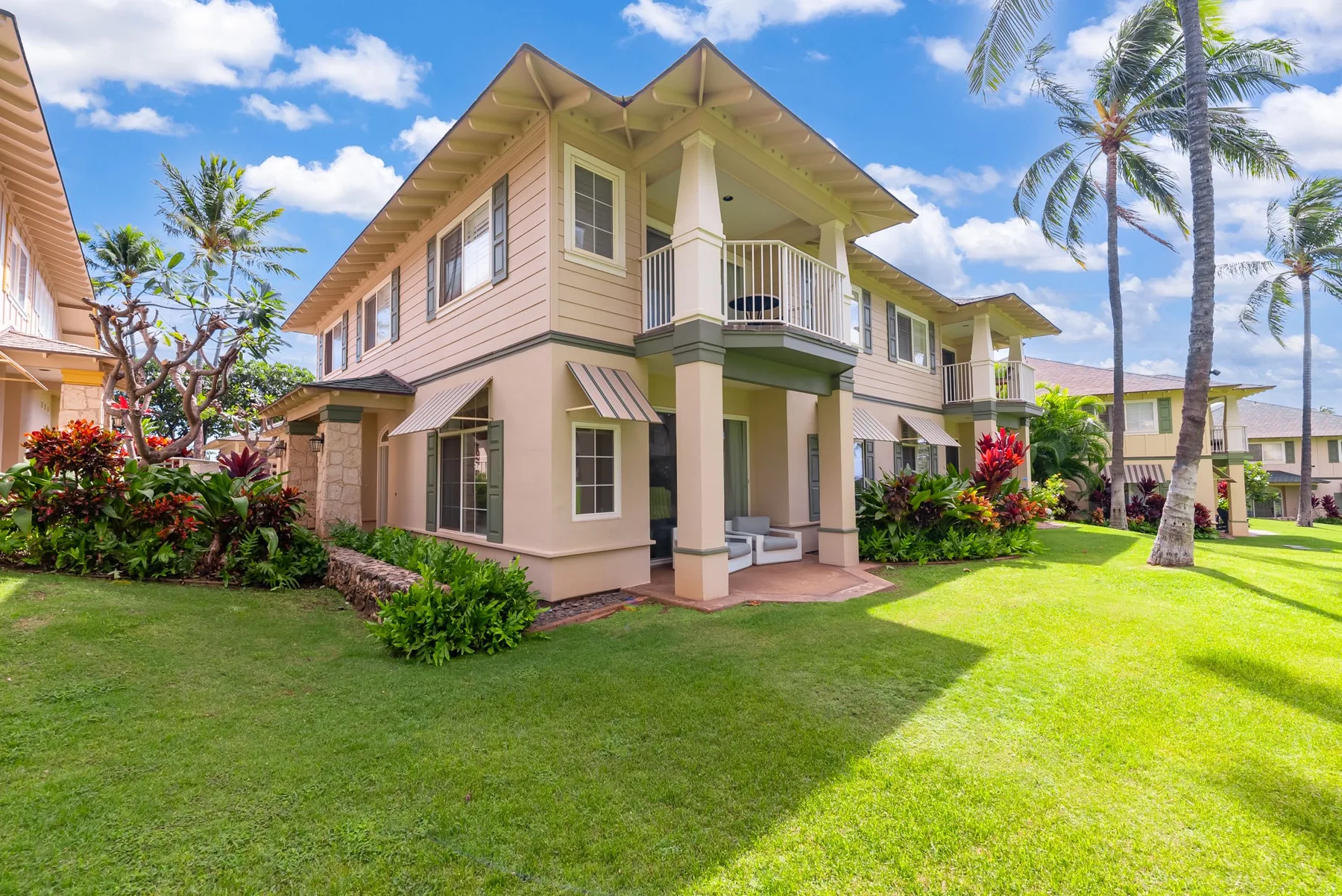 Una casa de dos plantas se encuentra junto a un césped verde y plantas tropicales.