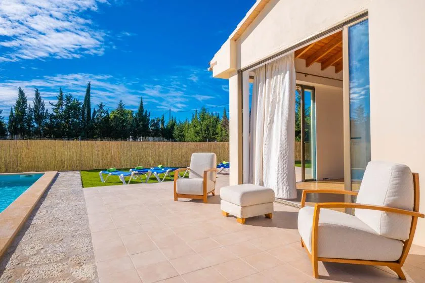 A sunny patio beside a swimming pool has cushioned seating and open sliding doors.