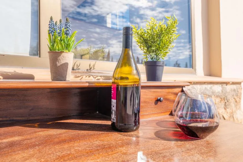 A bottle of wine and a glass sit on a sunlit wooden counter by the window.