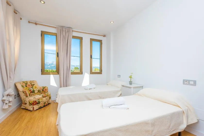 A bright bedroom with two beds, three windows, and a floral upholstered chair.