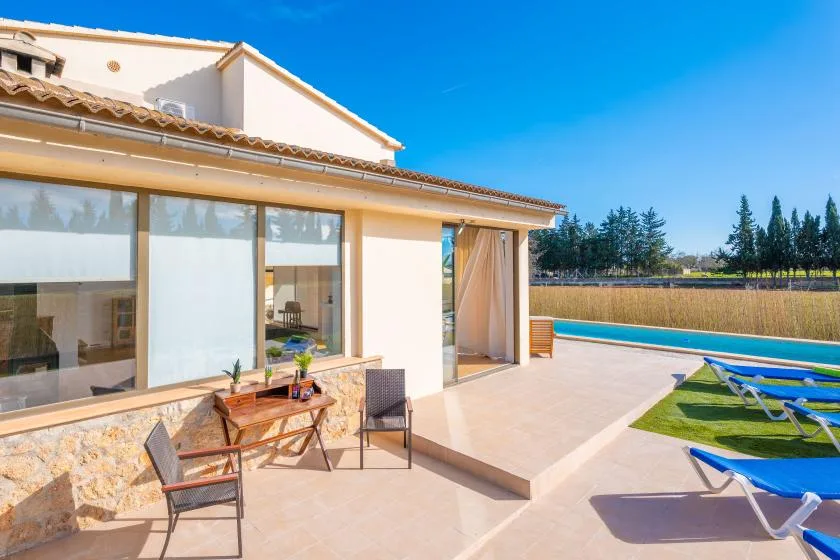 A sunny patio beside a modern house overlooks a swimming pool and lawn.