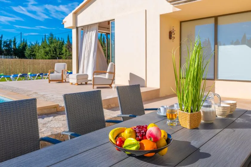A patio dining table with fruit overlooks a pool beside a modern house.