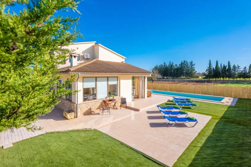 A house opens onto a paved patio with a swimming pool and lounge seating.