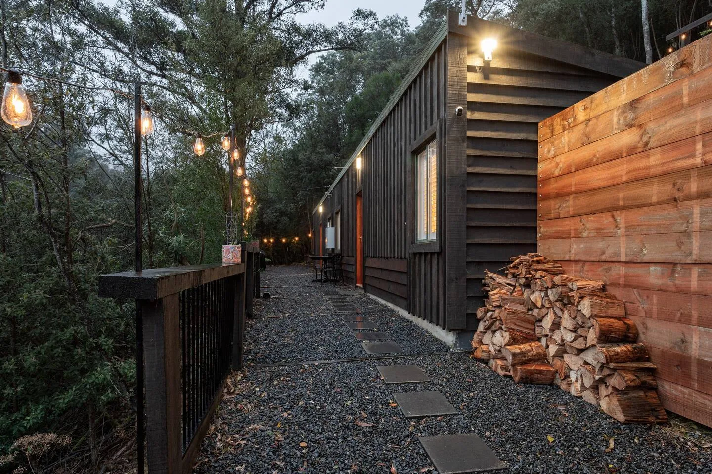 A gravel walkway runs beside a wood-sided building, with stacked firewood along the wall.
