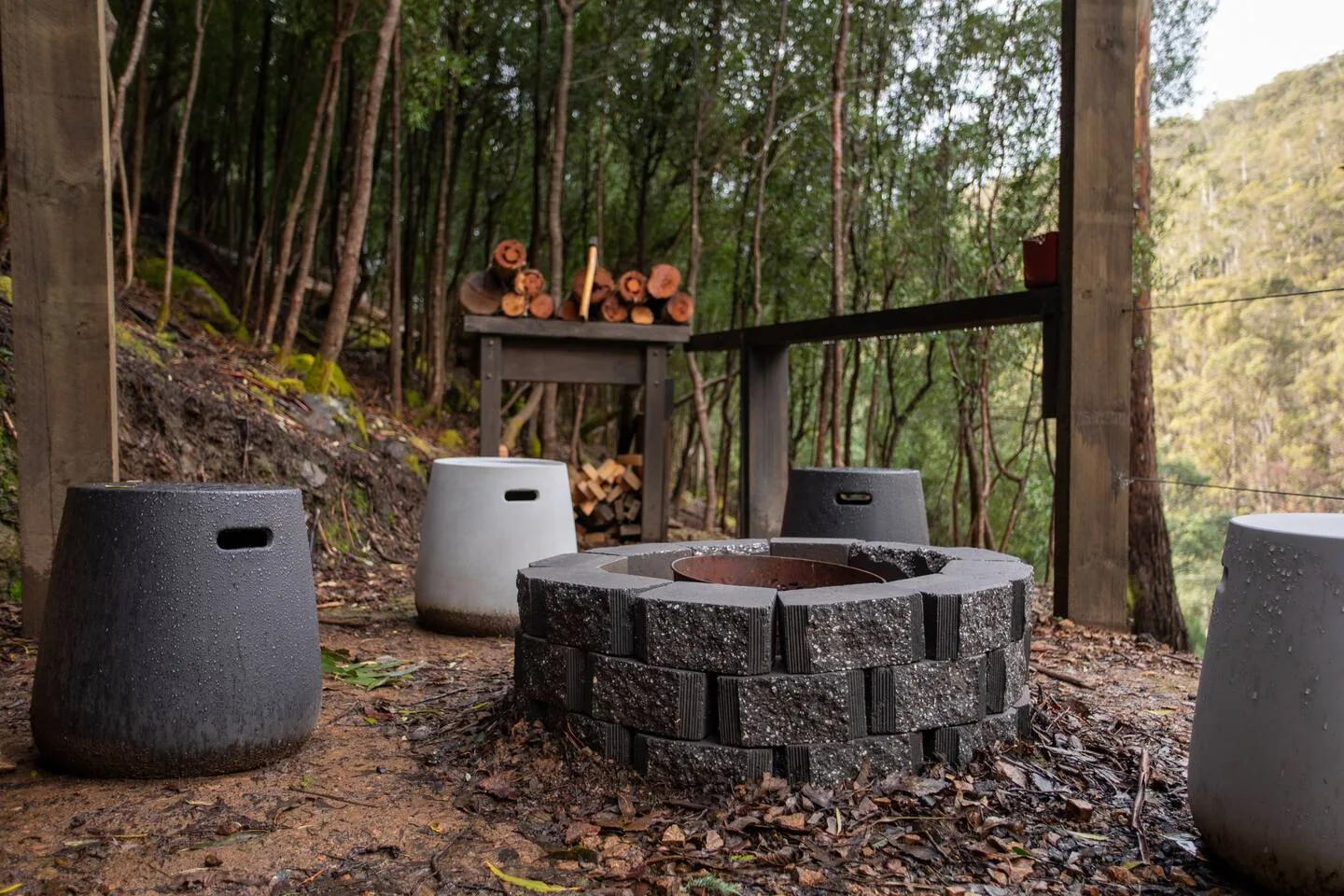 An outdoor fire pit with seating sits among trees beside a wood storage rack.
