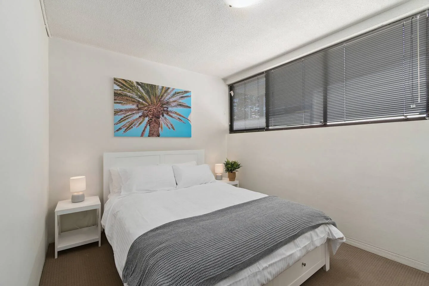 A bedroom with a bed, gray bedding, and palm tree artwork on the wall.