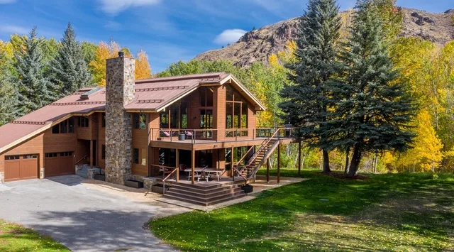 Una amplia casa de madera con chimenea de piedra se encuentra entre árboles bajo una montaña.