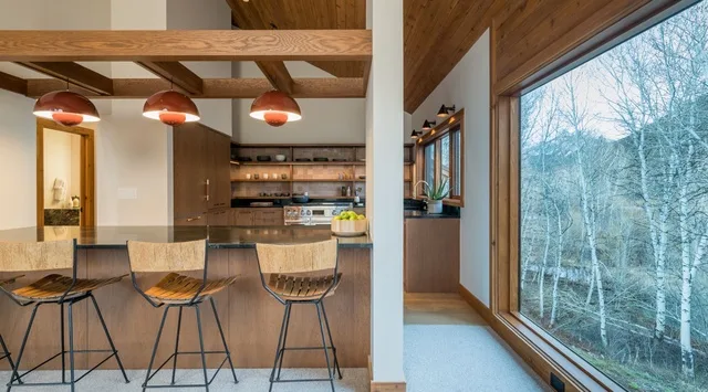 Una cocina y comedor con asientos en la barra y grandes ventanales con vistas a los árboles.