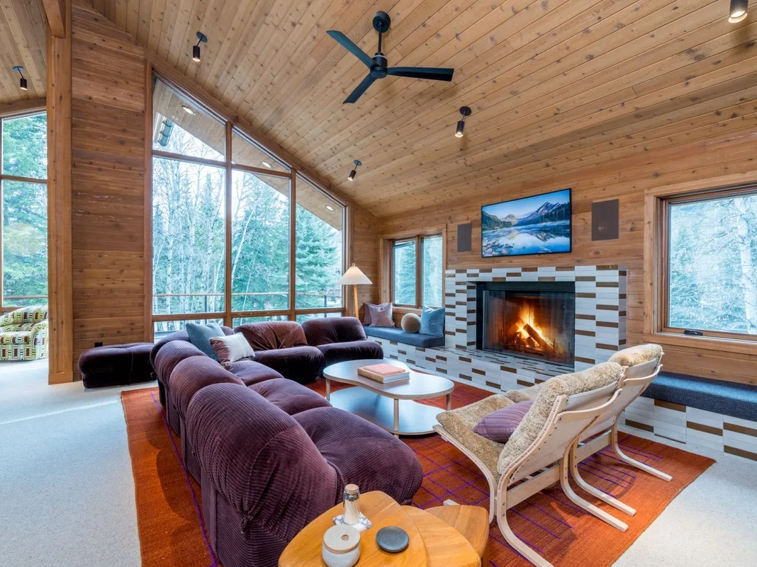 Una sala de estar revestida de madera tiene grandes ventanas, asientos y una chimenea.