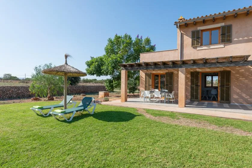 A house opens onto a grassy outdoor patio with seating and a shade umbrella.
