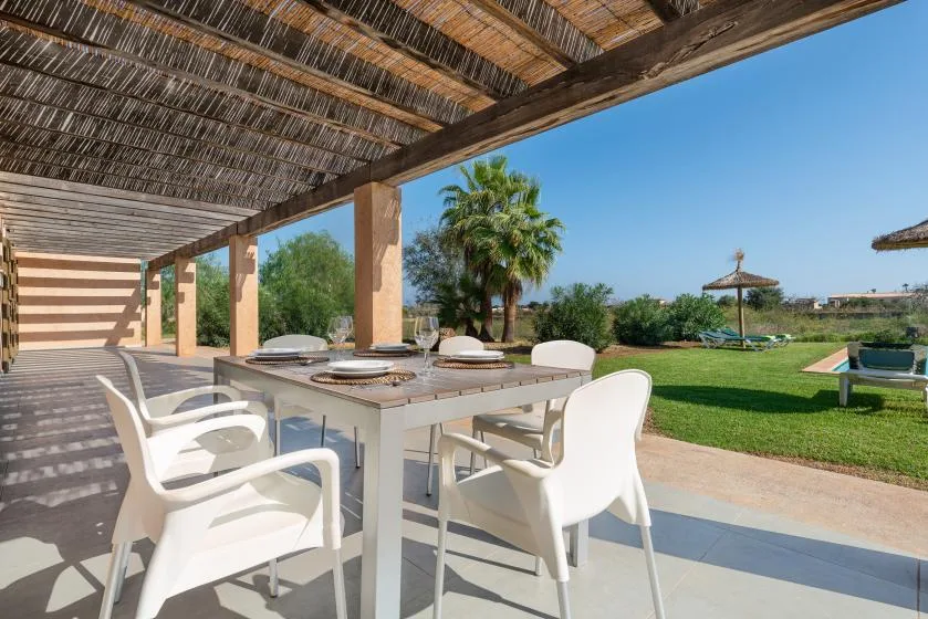 A covered outdoor dining area with a table and white seating overlooks a garden.