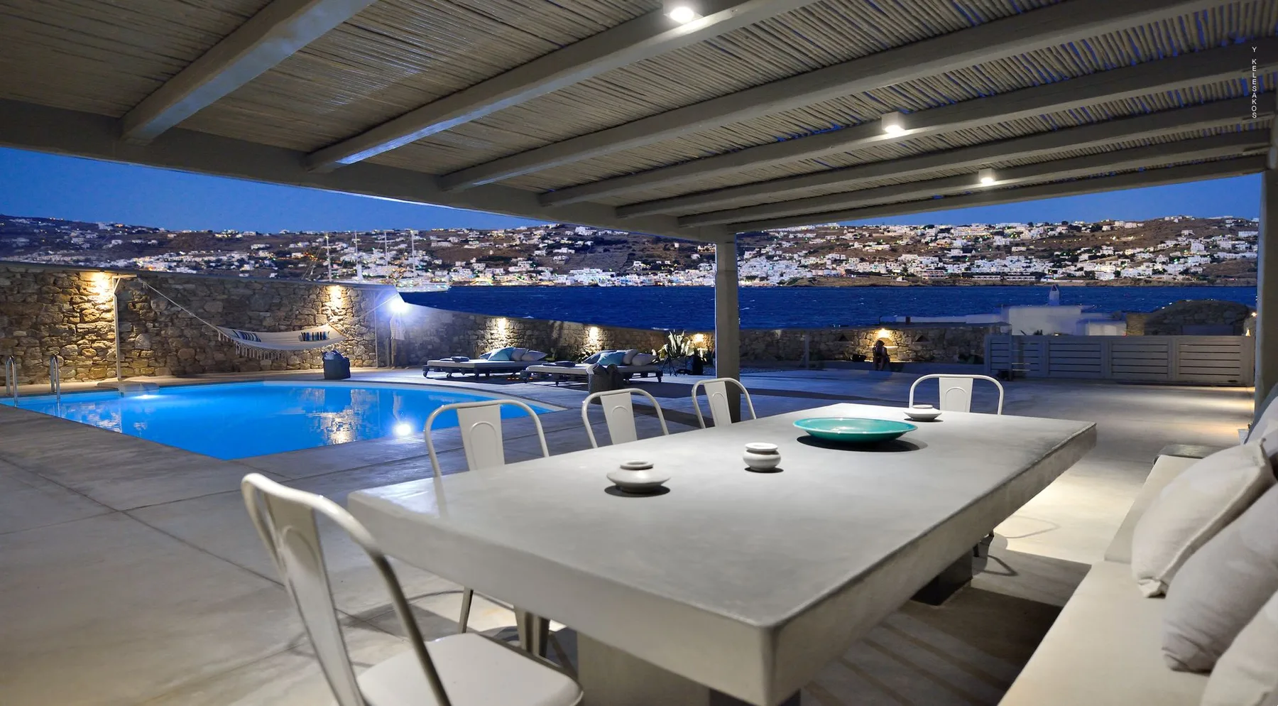 A covered outdoor dining area overlooks a swimming pool and coastal hillside.