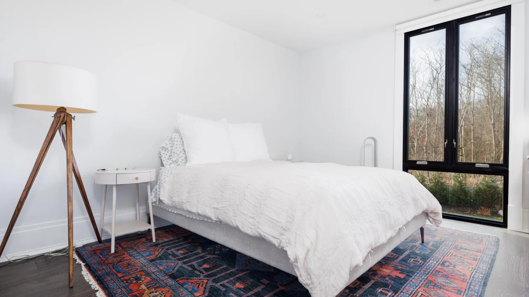 A bright bedroom with a white bed beside a large black-framed window.