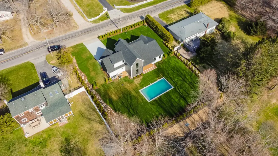 An aerial view shows several houses and a swimming pool amid green lawns and trees.