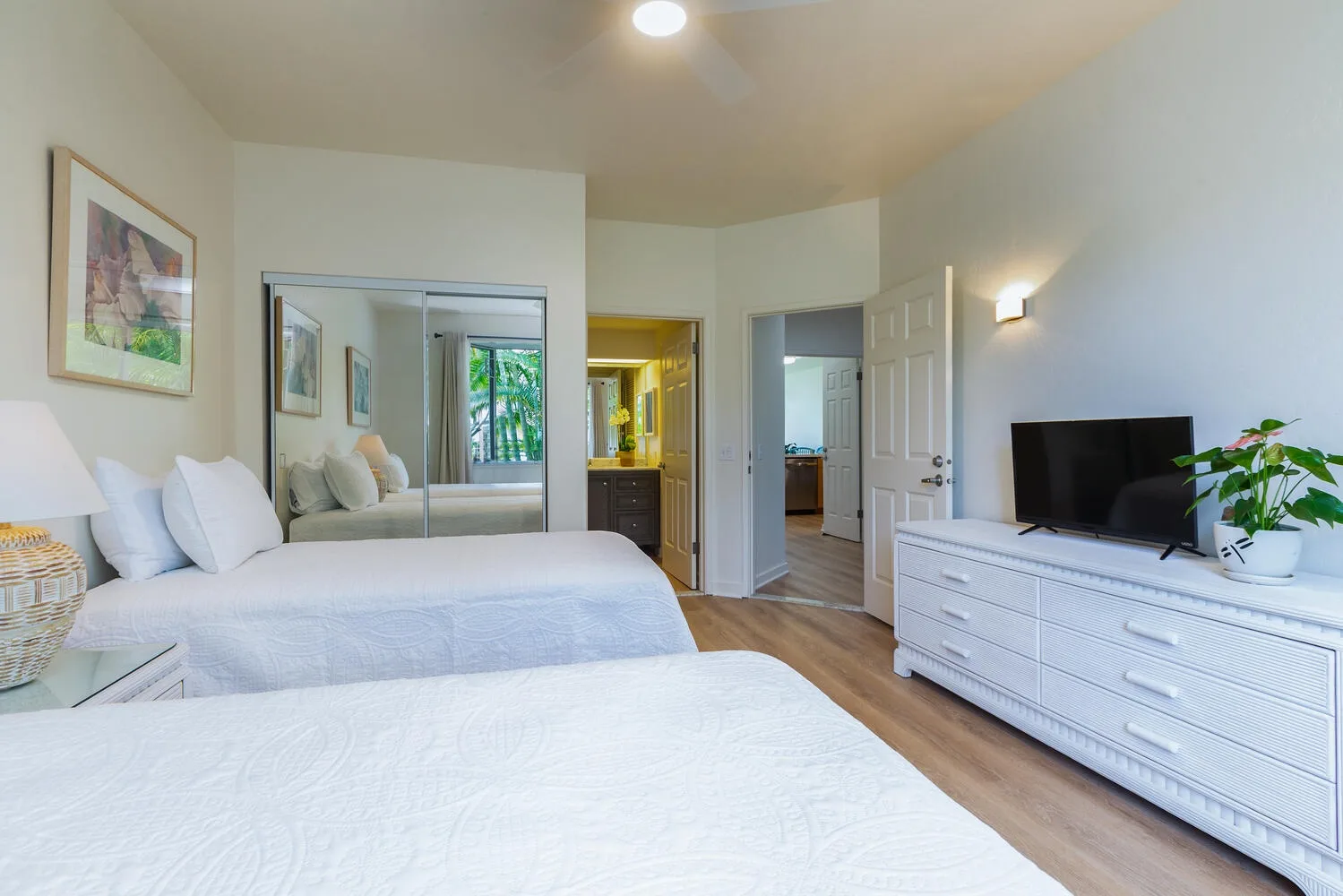 Un dormitorio luminoso con ropa de cama blanca, puertas de armario con espejo y una cómoda con televisor.