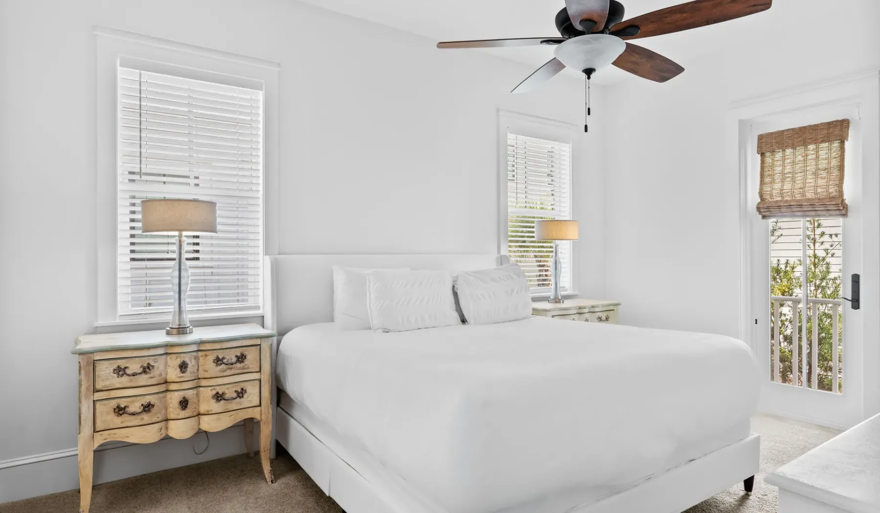 Un dormitorio luminoso con una cama blanca, mesitas de noche y ventanas con persianas.