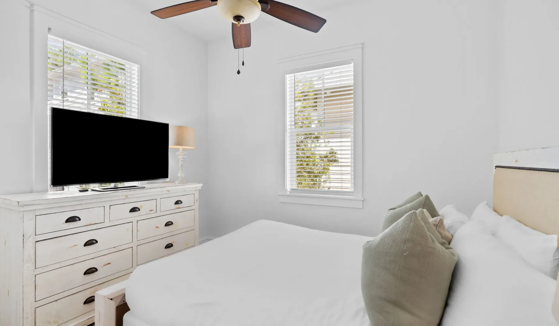 Un dormitorio luminoso con ropa de cama blanca y un televisor sobre una cómoda.