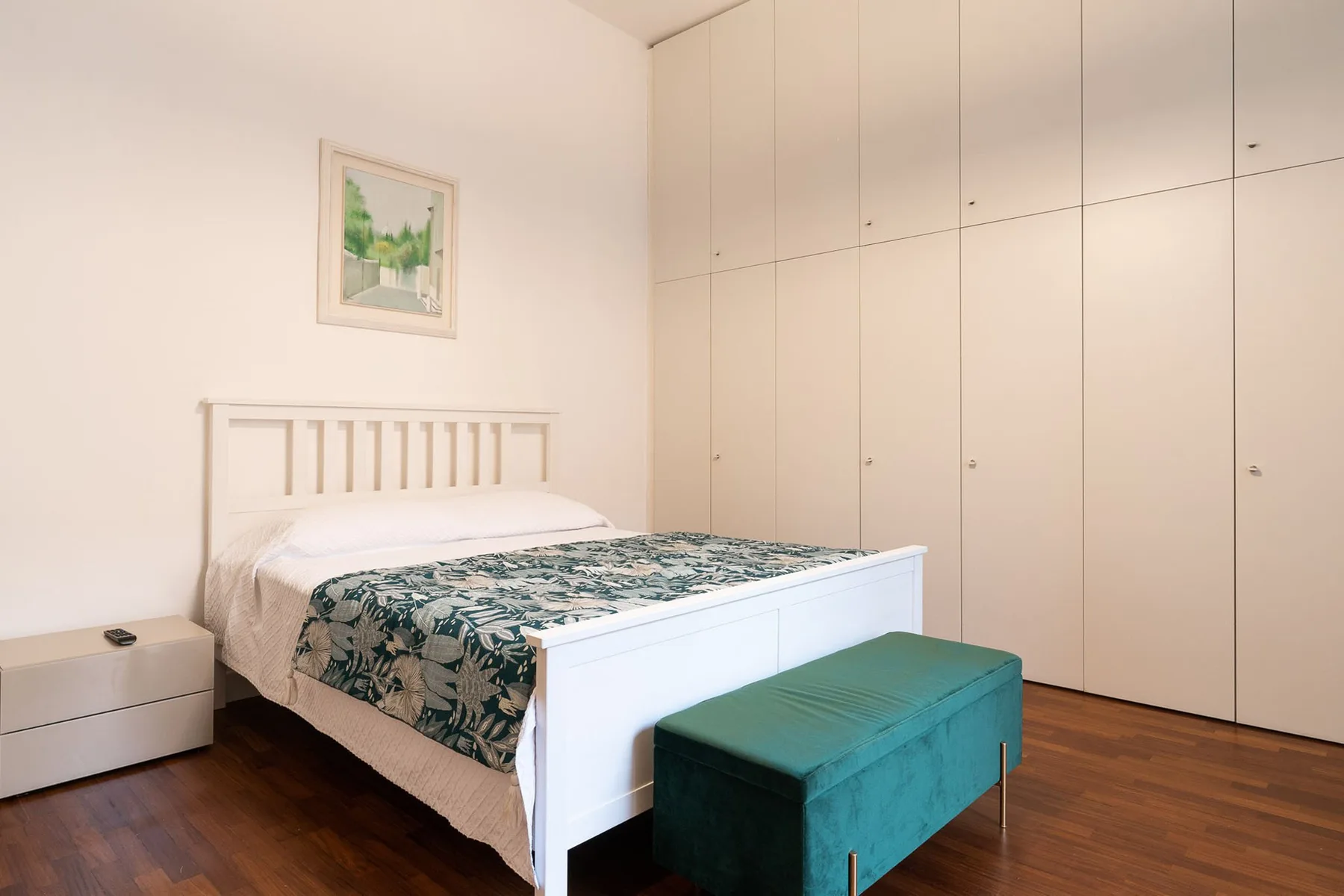 Un dormitorio con una cama blanca, ropa de cama estampada y un banco verde azulado al pie.