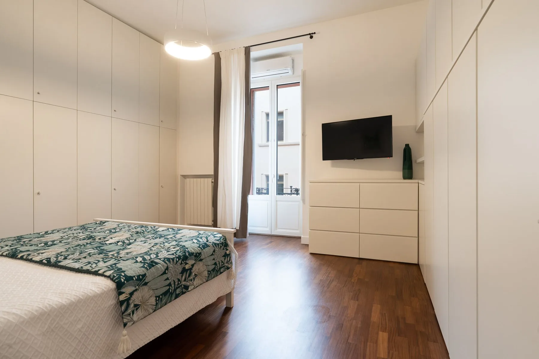 Un dormitorio con una cama estampada, un televisor en la pared y armarios empotrados altos.