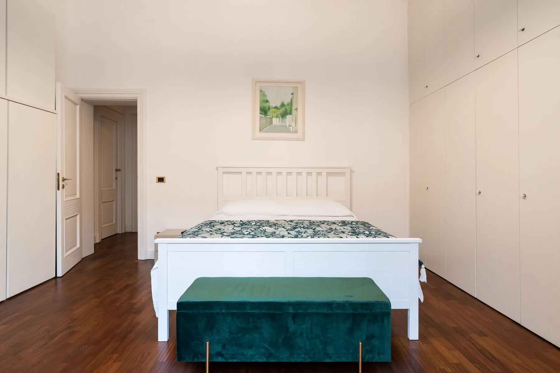 Un dormitorio luminoso con una cama blanca, un banco verde y armarios empotrados.