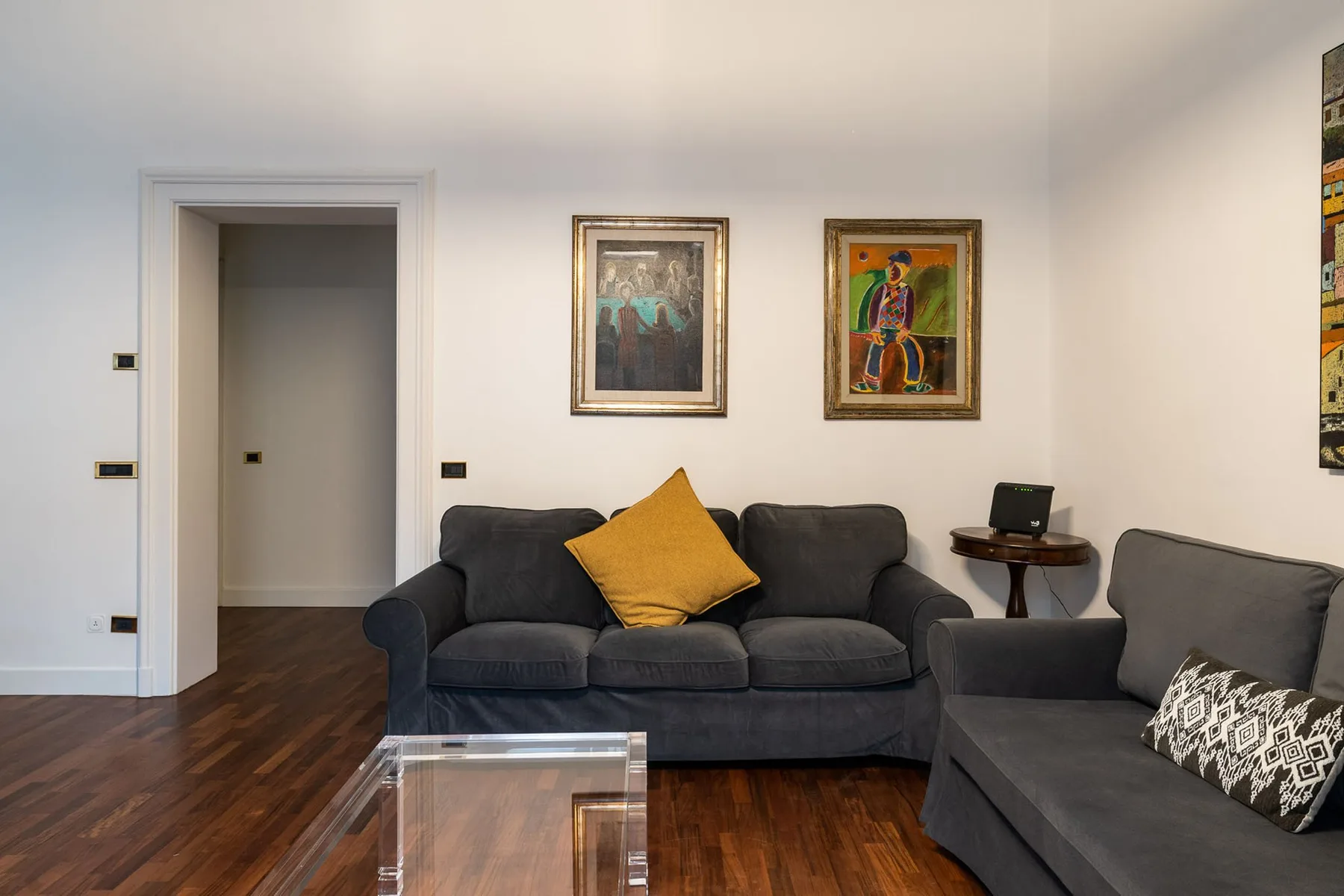 Una sala de estar con asientos oscuros, cuadros enmarcados y una mesa baja de cristal.
