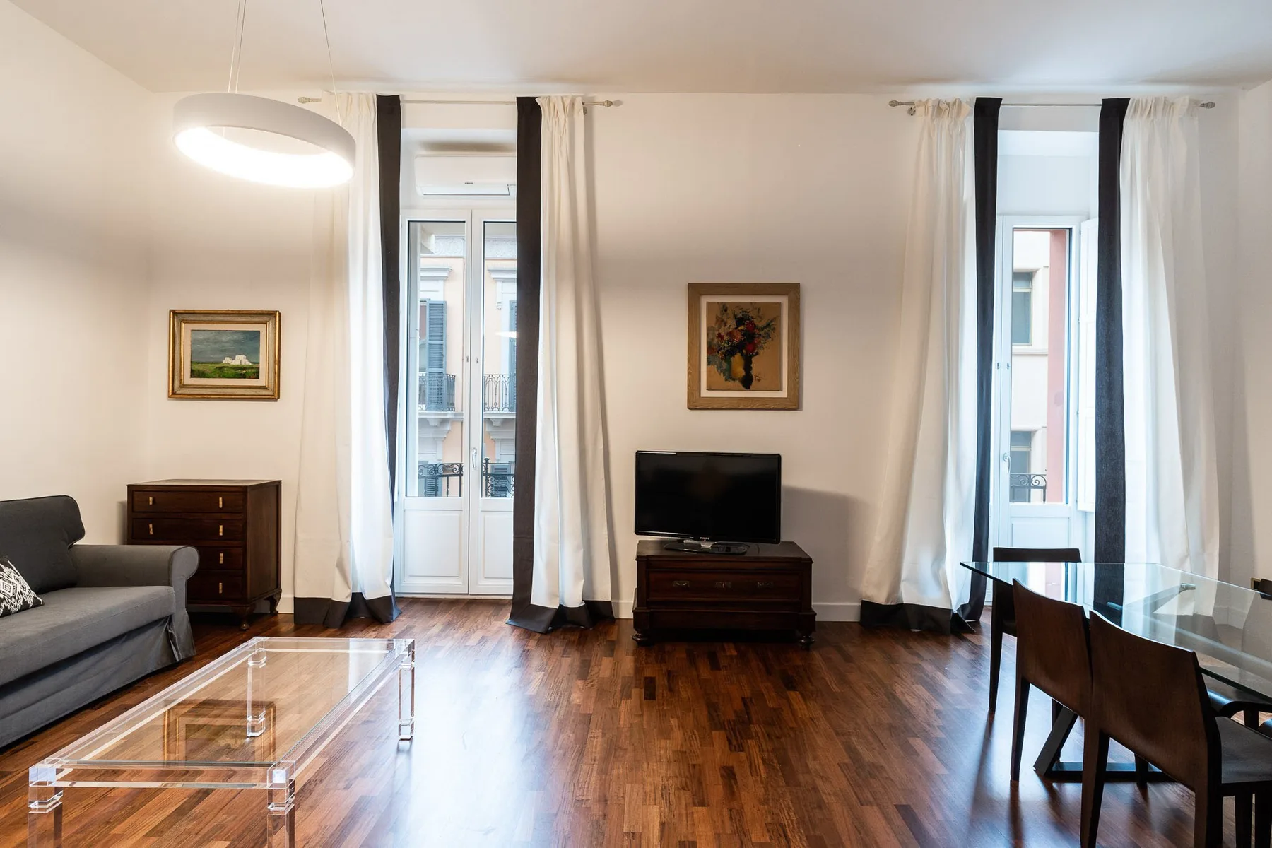 Una sala de estar con televisor, asientos y una mesa de comedor junto a ventanas altas.