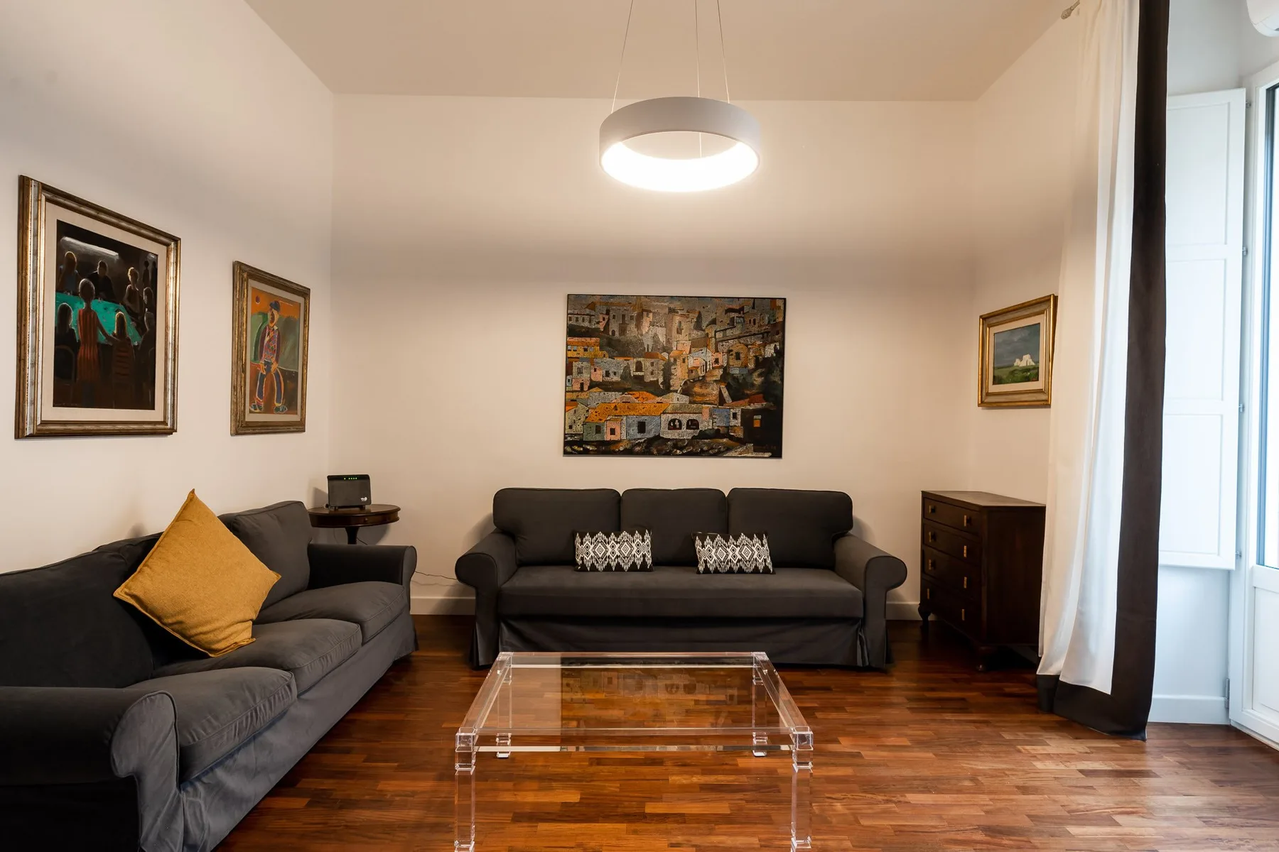 Una sala de estar con asientos oscuros, una mesa de centro transparente y cuadros enmarcados.