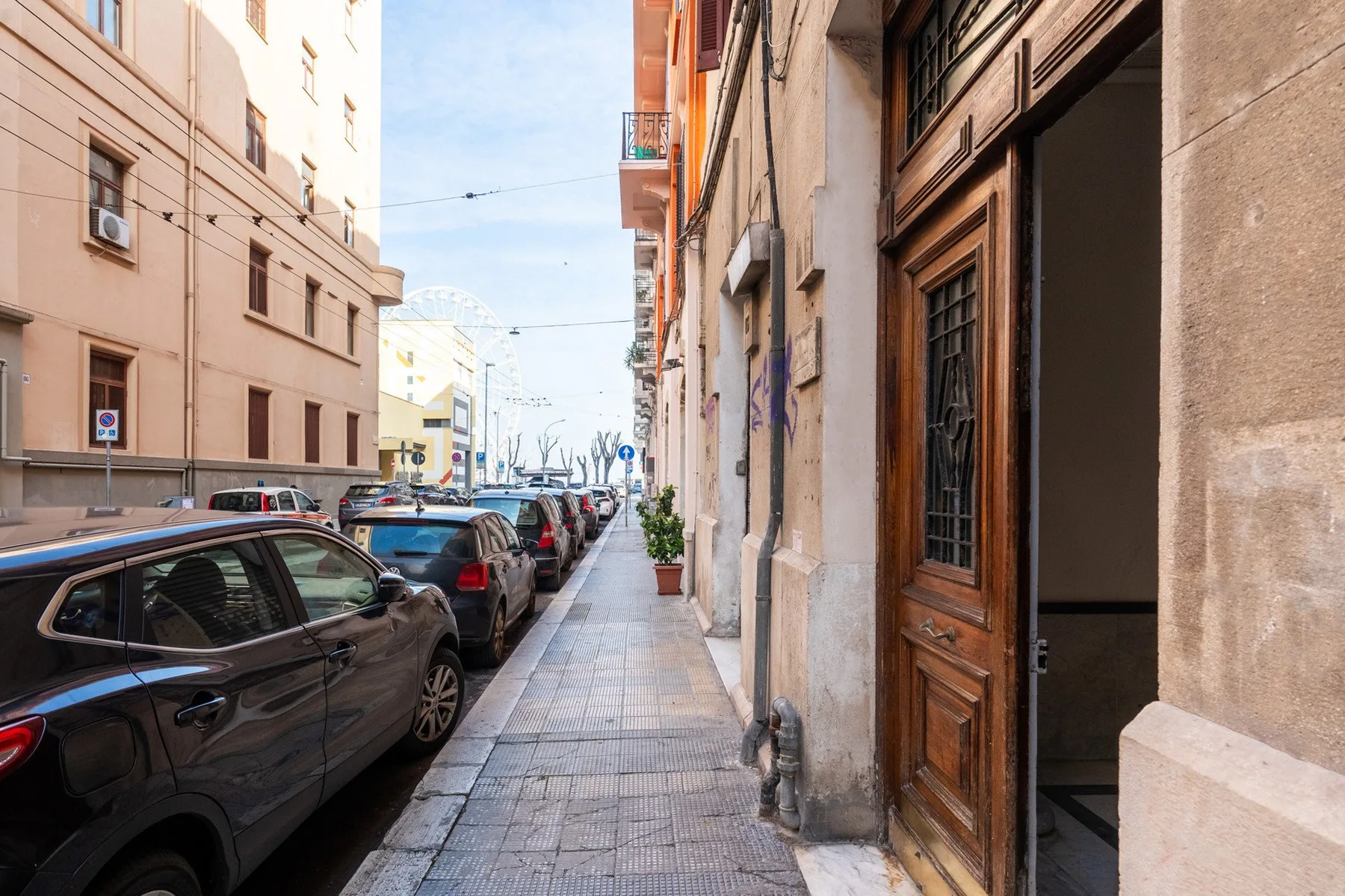 Una acera estrecha pasa entre coches estacionados y una puerta de madera abierta.