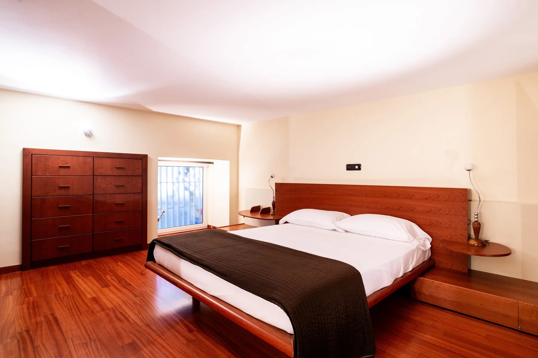 Un dormitorio con una cama grande, una cómoda oscura y suelo de madera pulida.