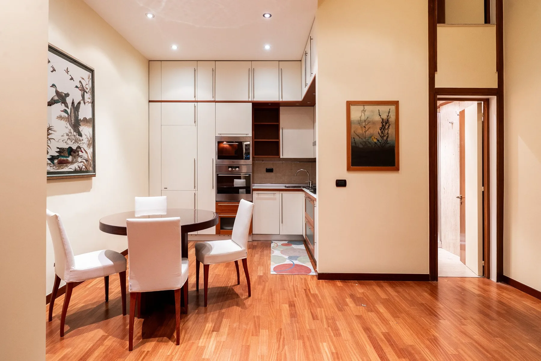 Un comedor se abre a una cocina compacta, con una mesa redonda y cuadros en la pared.
