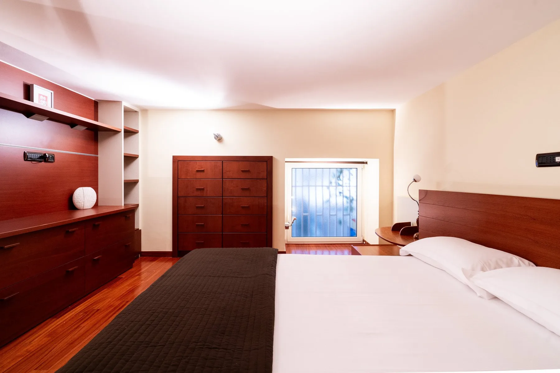 Un dormitorio con una cama blanca, una cómoda de madera oscura y una ventana.